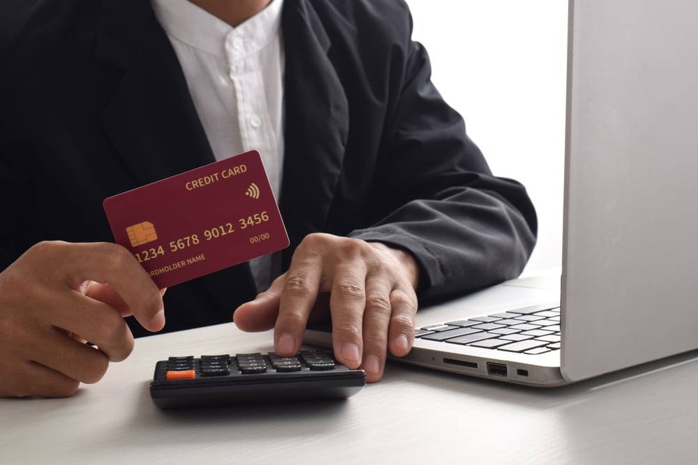 Businessperson in a suit using a business bank account; they are holding a credit card in one hand, typing on a calculator with the other, and working on a laptop at a desk.