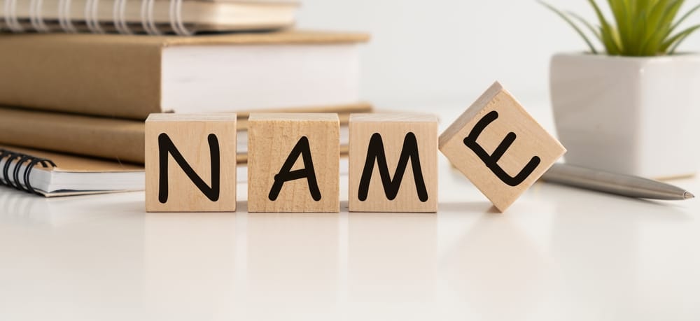 Wooden blocks spelling out the word "NAME" on a desk with notebooks and a pen in the background, suggesting the step to choose company name when starting a new business.